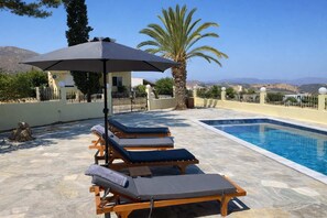 A poolside area with lounge chairs and a large umbrella, surrounded by a stone patio and a view of distant hills. - Creta Leisure Escape Private Pool Garden Oasis (Faistos)