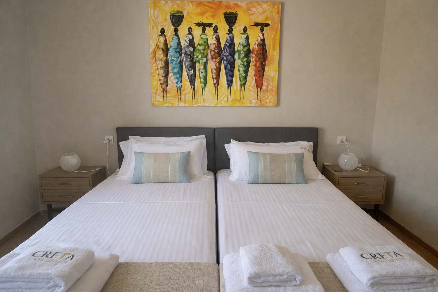 Une chambre double avec deux lits, chacun agrémenté de draps blancs et d’oreillers à rayures, un tableau au-dessus des lits et une petite table de chevet avec une lampe.