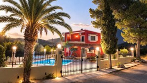 Eine rote Villa mit Swimmingpool, umgeben von einem Zaun und Palmen, mit einer Bergkulisse im Hintergrund.