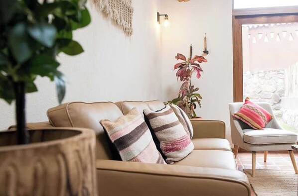 A beige leather sofa with striped cushions, a wooden side table with a plant, and a white armchair with a colorful cushion. - w Serene 2BR Riverside Villa w Nature Views (Calca)