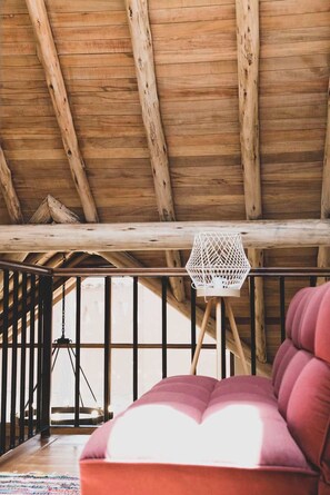 Un plafond en bois avec des poutres apparentes, un luminaire suspendu blanc et un coin salon rouge avec des coussins.