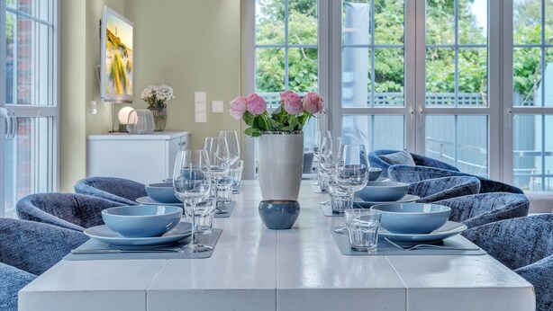Un coin repas avec une table dressée pour six personnes, des chaises bleues, un vase rempli de fleurs roses et un tableau accroché au mur.