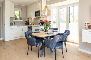 A modern kitchen with white cabinets, a wooden countertop, and a dining area with a round table and blue chairs. - Exclusive thatched house in Wenningstedt with sauna, garden and modern furnishings (Wenningstedt)