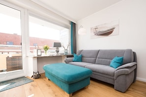 A modern living room with a grey sofa, teal ottoman, and a window with a view of buildings. - Modern apartment with balcony and comfortable furnishings in Westerland (Westerland)