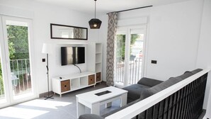 Una sala moderna con un sofá blanco, una mesa de centro, un televisor de pantalla plana y un ventanal con vistas a la vegetación.