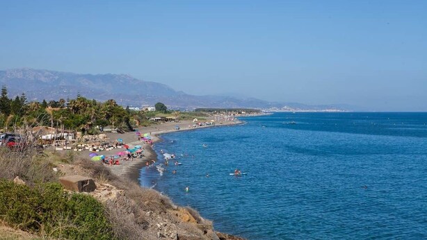 Una playa costera con aguas azules cristalinas, una orilla arenosa y montañas al fondo.