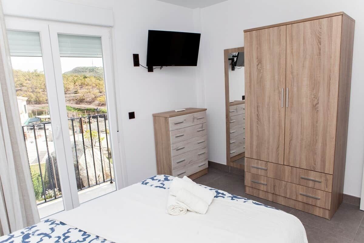 Un dormitorio con cama, un armario de madera, un televisor montado en la pared y un balcón con vistas a la vegetación.