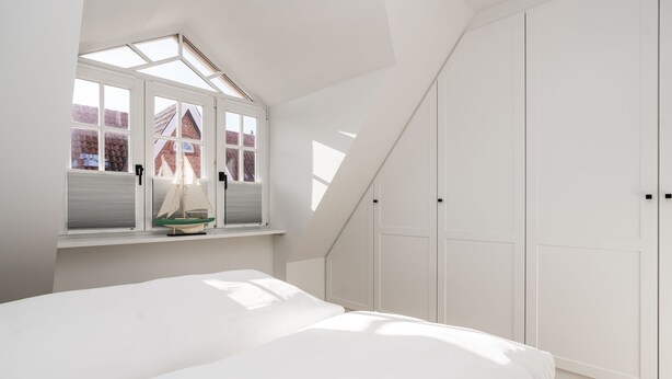 A bedroom with a sloped ceiling, a bed, a window with a sailboat decoration, and white wardrobes. - Haus Friesenperle: Comfortable vacation home with terrace and garden in Westerland (Westerland)