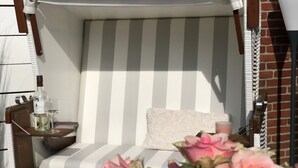 En mysig, randig bänk med kuddar och en prydnadskudde, ett litet bord med en flaska och ett glas, och rosa blommor i förgrunden.