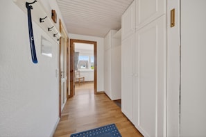 A hallway with white walls, wooden flooring, and a blue rug. There are white doors, a wooden-framed window, and a chair in the background. - Comfortable vacation apartment with sauna, garden and terrace in Tinnum on Sylt (Tinnum)