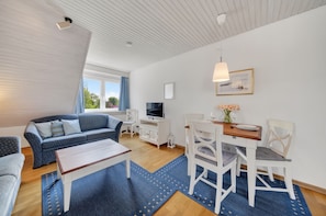 A modern living room with a sofa, dining table, and chairs, a TV, and a window with curtains. - Comfortable vacation apartment with sauna, garden and terrace in Tinnum on Sylt (Tinnum)