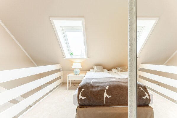 A bedroom with a sloped ceiling, two windows, a bedside table with a lamp, a bed with a brown blanket, and a white railing. - Beautiful vacation apartment with sauna, indoor pool and fitness room in Keitum (Keitum)