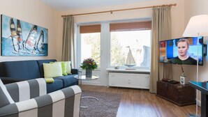 En stue med sofa, fjernsyn, et vindue med gardiner, et billede og en radiator.