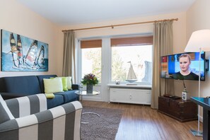 A living room with a sofa, a television, a window with curtains, a painting, and a radiator. - Modern vacation apartment with terrace and garden in Wenningstedt (Wenningstedt)