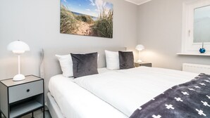 Et soveværelse med en seng, to sengeborde, et natbord, et vindue og en vægdekoration med et strandmotiv.