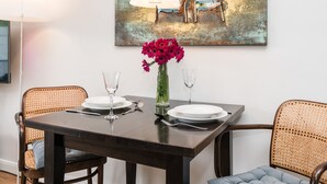 Spisebord dækket med tallerkener, bestik og en blomstervase, omgivet af fletstole, med et maleri med strandmotiv over.