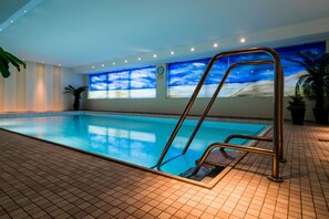 An indoor swimming pool with clear blue water, a metal ladder, tiled floor, and a large window with a sky view. - Modern vacation apartment with sauna, solarium and heated indoor pool in Westerland (Westerland)