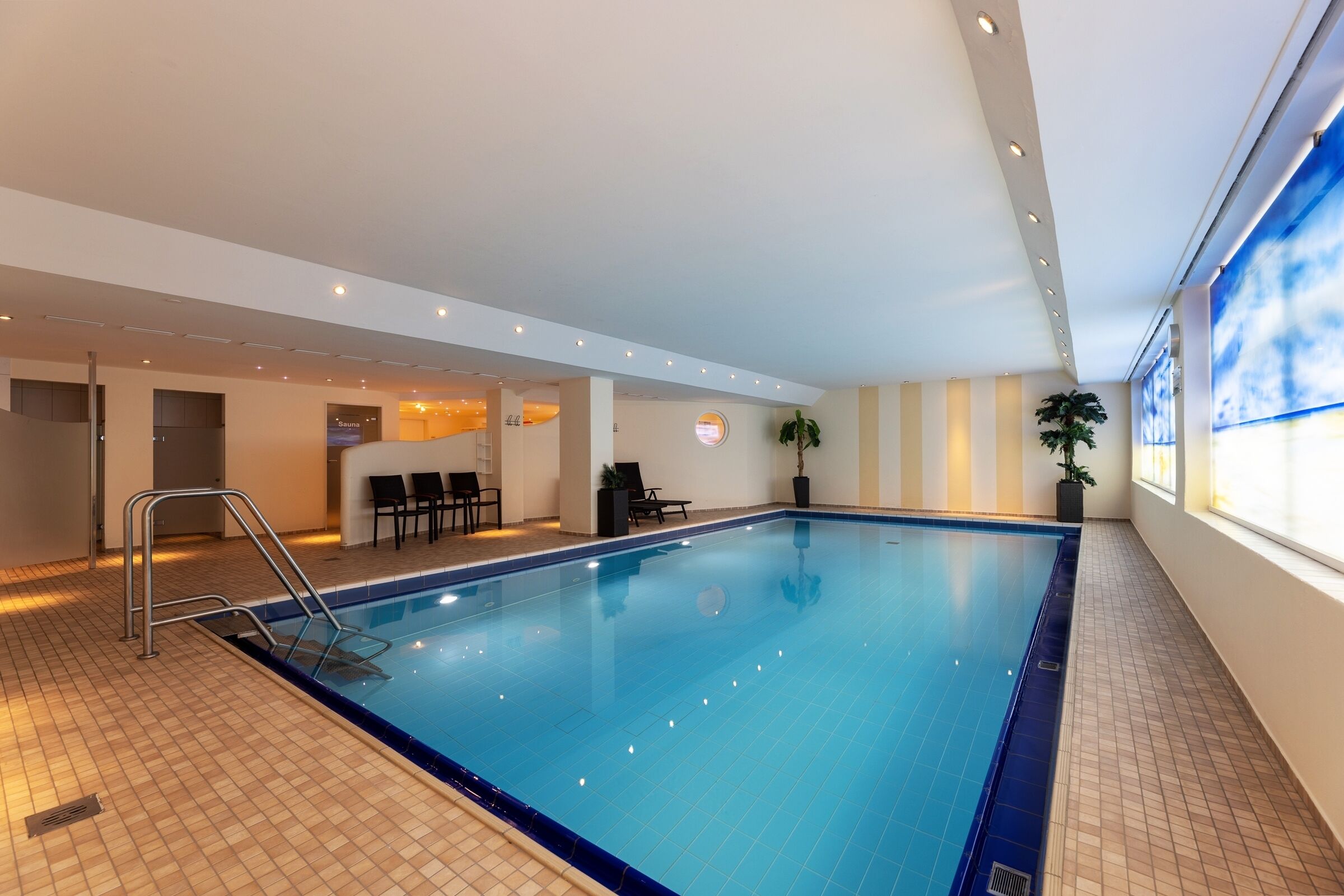 An indoor swimming pool with a tiled floor, a metal ladder, and a seating area with chairs and a table.