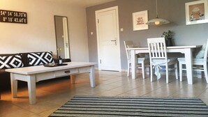 A living room with a white sofa, a white coffee table, a dining table with white chairs, a black and white rug, and a white door. - Comfortable vacation apartment with terrace in Westerland (Westerland)