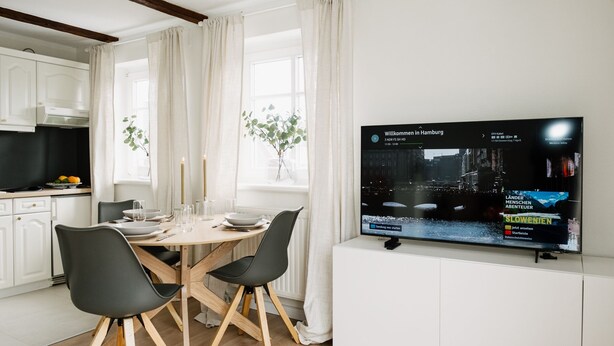 Eine moderne Küche mit weißen Schränken, einem Esstisch mit Stühlen und einem an der Wand befestigten Flachbildfernseher, der einen Nachrichtensender zeigt.