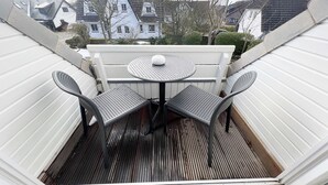 Ein überdachter Balkon mit rundem Tisch und zwei Stühlen, mit Blick auf eine Wohngegend mit Häusern und Bäumen.
