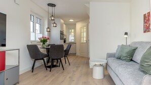 A modern living room with a grey sofa, a dining area with a table and chairs, and a kitchen in the background. - This beautiful and new end terraced house is centrally located in Westerland (Westerland)