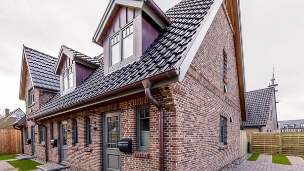A two-story brick house with a sloped roof, a front door, and a small front yard with a pathway and a wooden fence. - This beautiful and new end terraced house is centrally located in Westerland (Westerland)