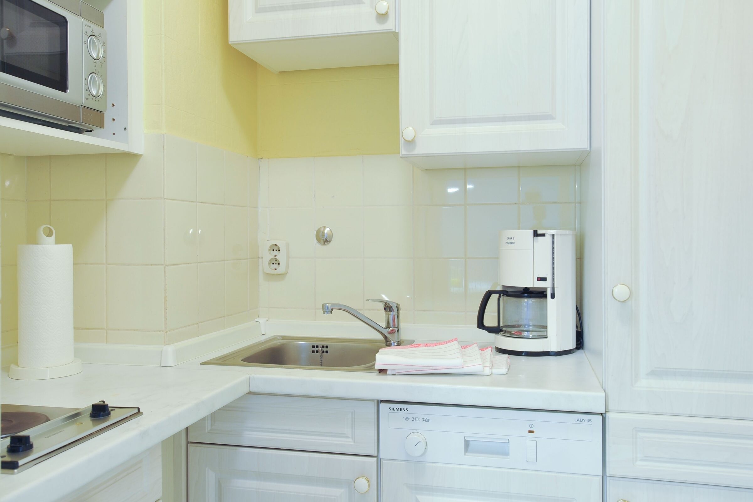 Una cocina con armarios blancos, microondas, lavabo, cafetera y lavavajillas Siemens.