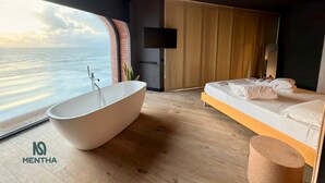 A modern bedroom with a large bathtub, a bed with white linens, a mounted TV, and a view of the ocean. - Mentha Mare e Spa (Salerno)