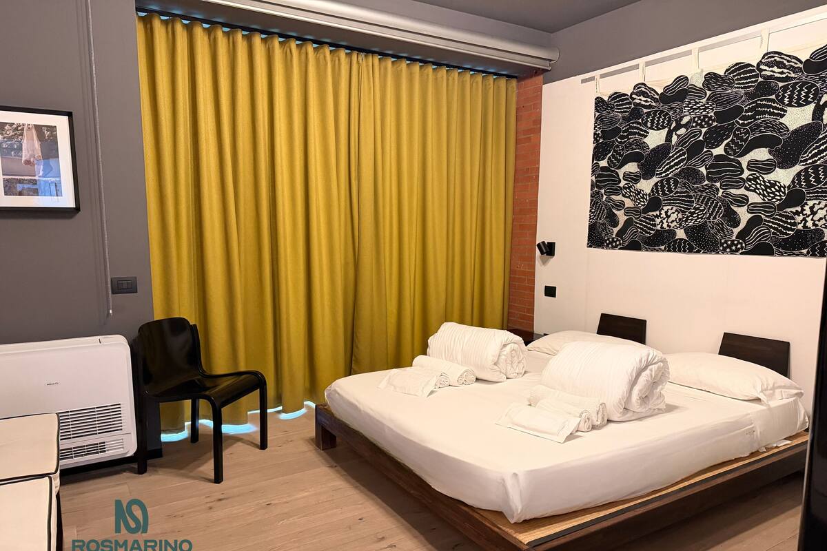Een hotelkamer met een bed, een stoel, een bureau en een groot raam met gele gordijnen.