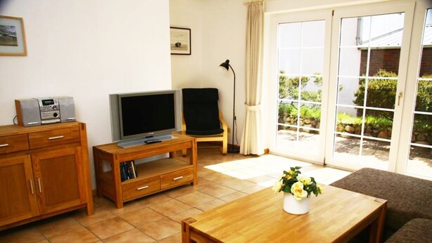 A living room with a wooden TV stand, a flat-screen TV, a sofa, a coffee table with a flower vase, and a sliding glass door leading to a garden. - Schmiedeweg Whg. 2 in Westerland - comfortable apartment with terrace and sauna (Westerland)