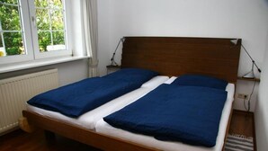 A double bed with a wooden headboard, a window with a view of greenery, and a bedside table with a lamp. - Schmiedeweg Whg. 2 in Westerland - comfortable apartment with terrace and sauna (Westerland)