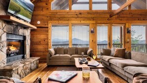 Interior acogedor de una cabaña con chimenea de piedra, paredes de madera y un ventanal con vistas a un paisaje montañoso.