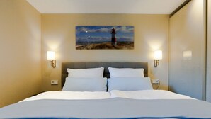 Hotellihuone, jossa on suuri sänky, kaksi yöpöytävalaisinta, sängynpääty ja seinällä oleva maalaus, jossa on kuvattuna majakka.