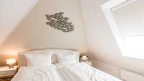 Ein Schlafzimmer mit einem ordentlich bezogenen Bett, einem Nachttisch mit Lampe, einem Wandbild und einem Fenster.