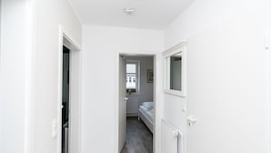 Ein schmaler Flur mit weißen Wänden und Fliesenboden führt zu einem Schlafzimmer mit Bett und Fenster.