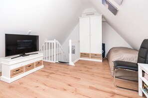 Une pièce moderne et bien éclairée, avec un téléviseur posé sur un meuble blanc, un lit avec un matelas beige, une chaise noire et une armoire blanche.
