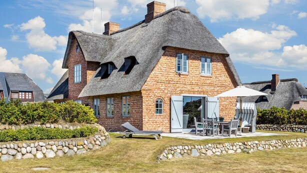 Ein Haus mit einem Reetdach, Ziegelwänden und einem Steinweg, der zu einer Terrasse mit Gartenmöbeln führt.