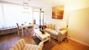 A living room with a sofa, a coffee table with fruit, a dining table with chairs, a floor lamp, and a painting on the wall. - Modern apartment in Westerland with balcony, parking and WiFi (Westerland)