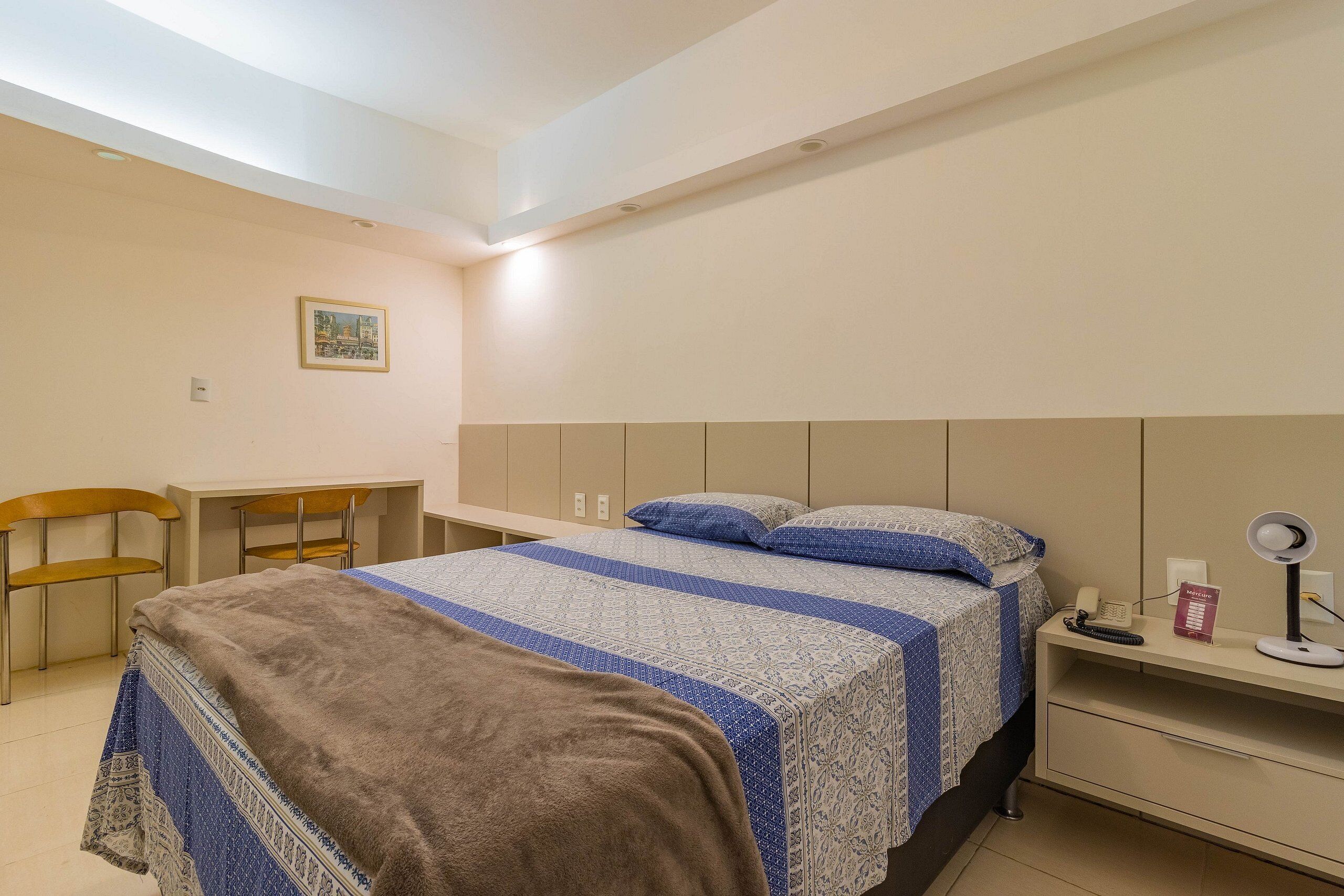 A bedroom with a bed, a desk, a chair, and a nightstand with a lamp and phone.