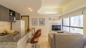 Una cocina moderna con encimeras de granito, microondas integrado y refrigerador. En la sala de estar, hay dos taburetes de bar y un sofá gris.