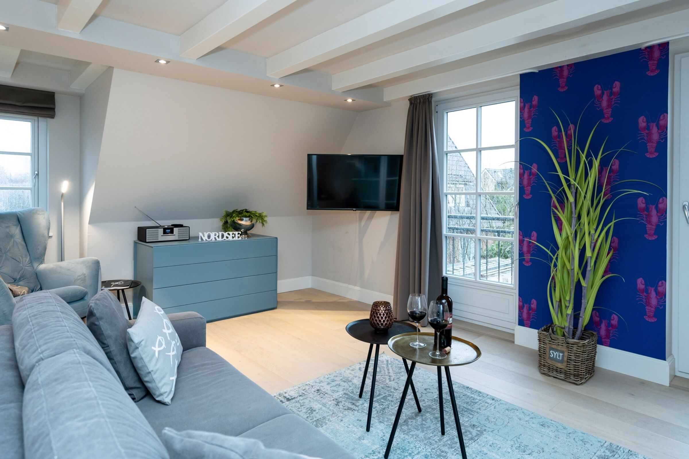 A modern living room with a grey sofa, a blue cabinet, a flat-screen TV, and a large window with curtains.