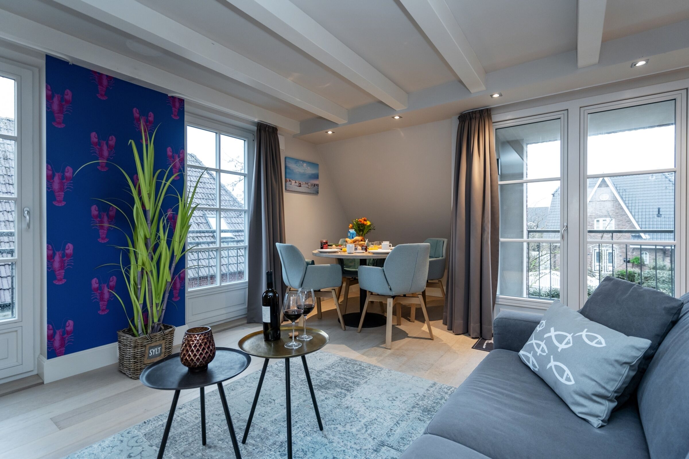 Un salon moderne avec un canapé gris, un coin repas avec une table dressée pour deux personnes, et de grandes fenêtres offrant une vue sur la ville.
