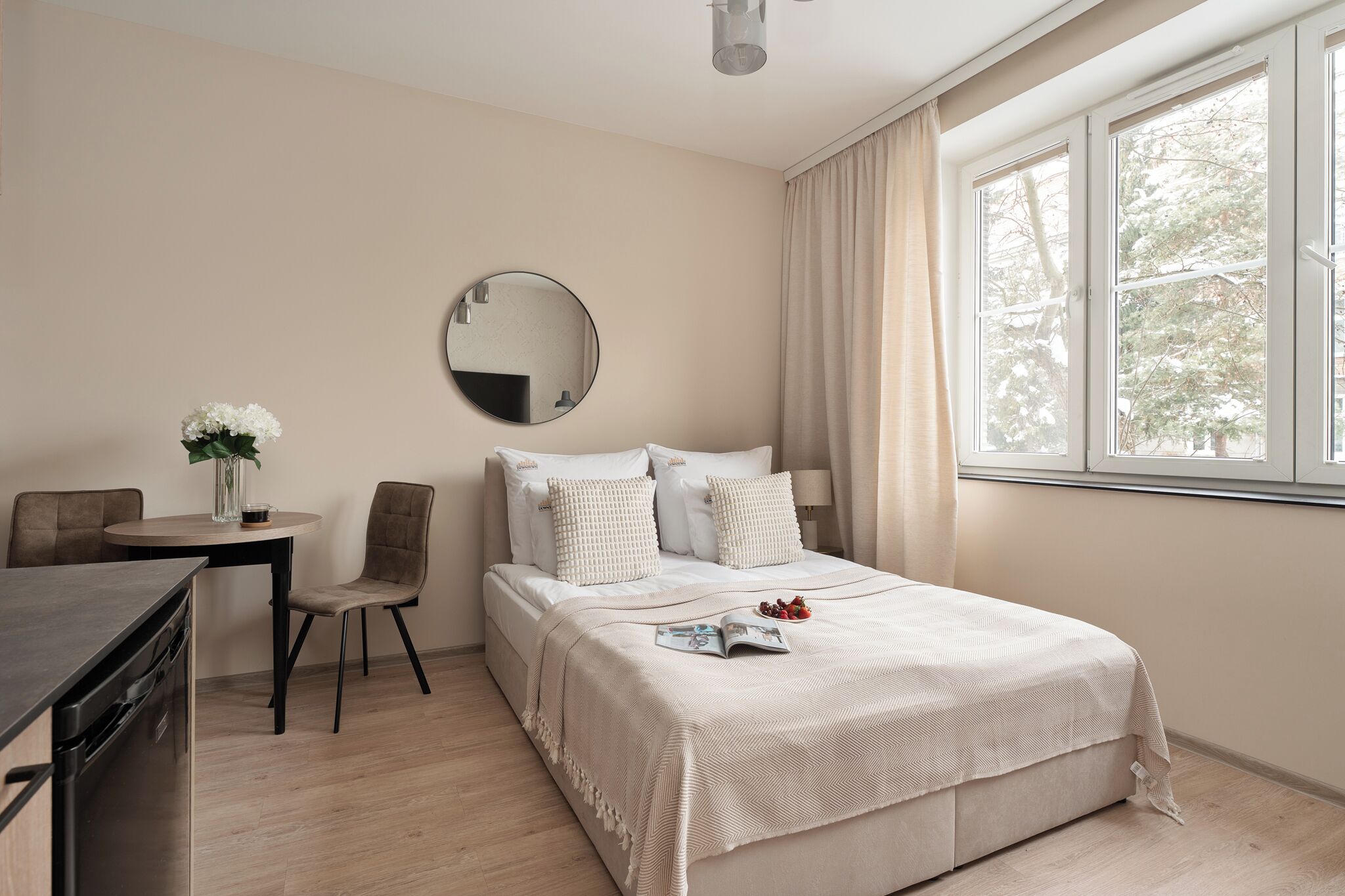 A bedroom with a bed, a bedside table, a chair, a round mirror, and a window with curtains.