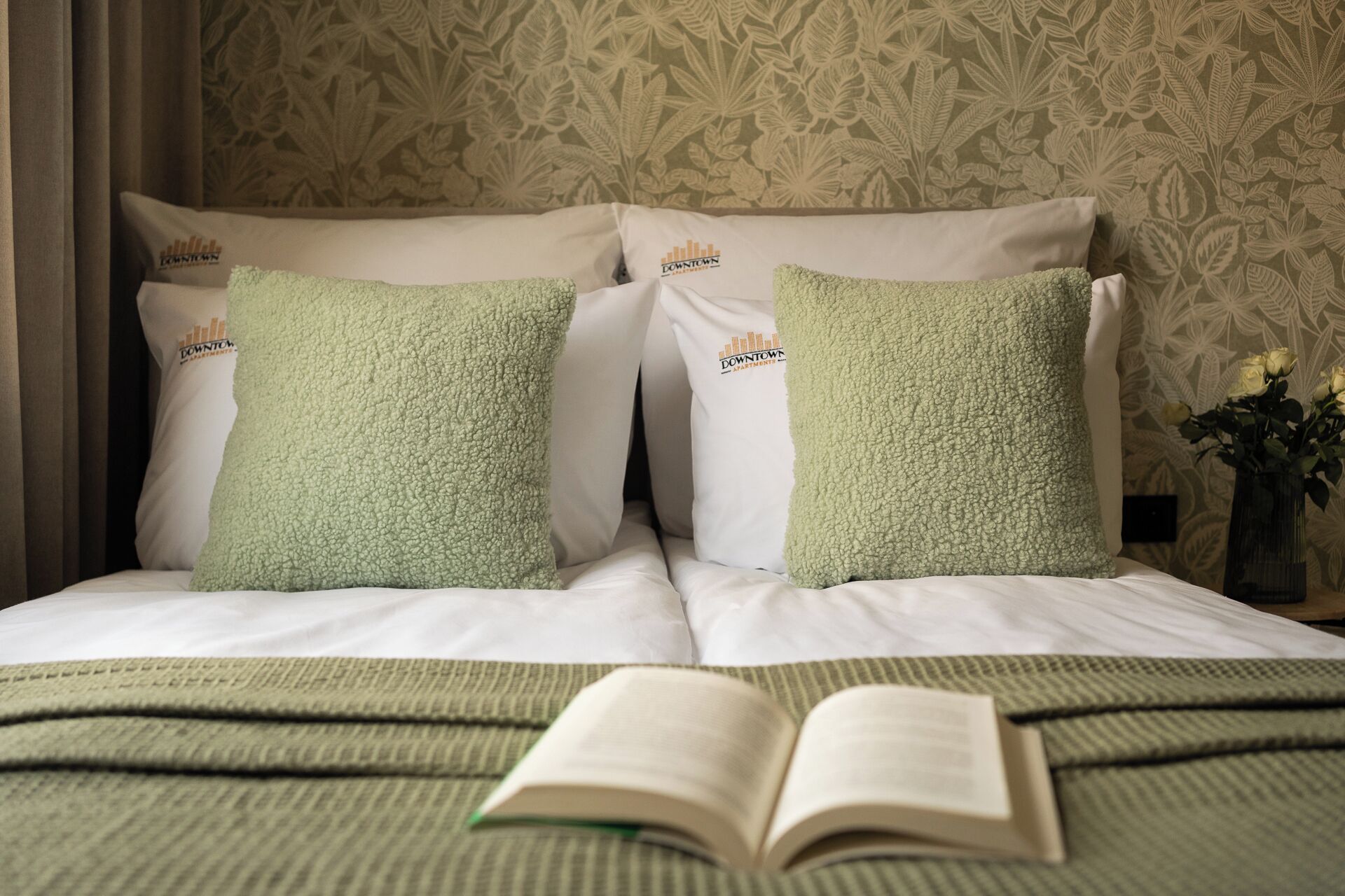 Una habitación de hotel con una cama, dos almohadas, un libro y un jarrón con flores.