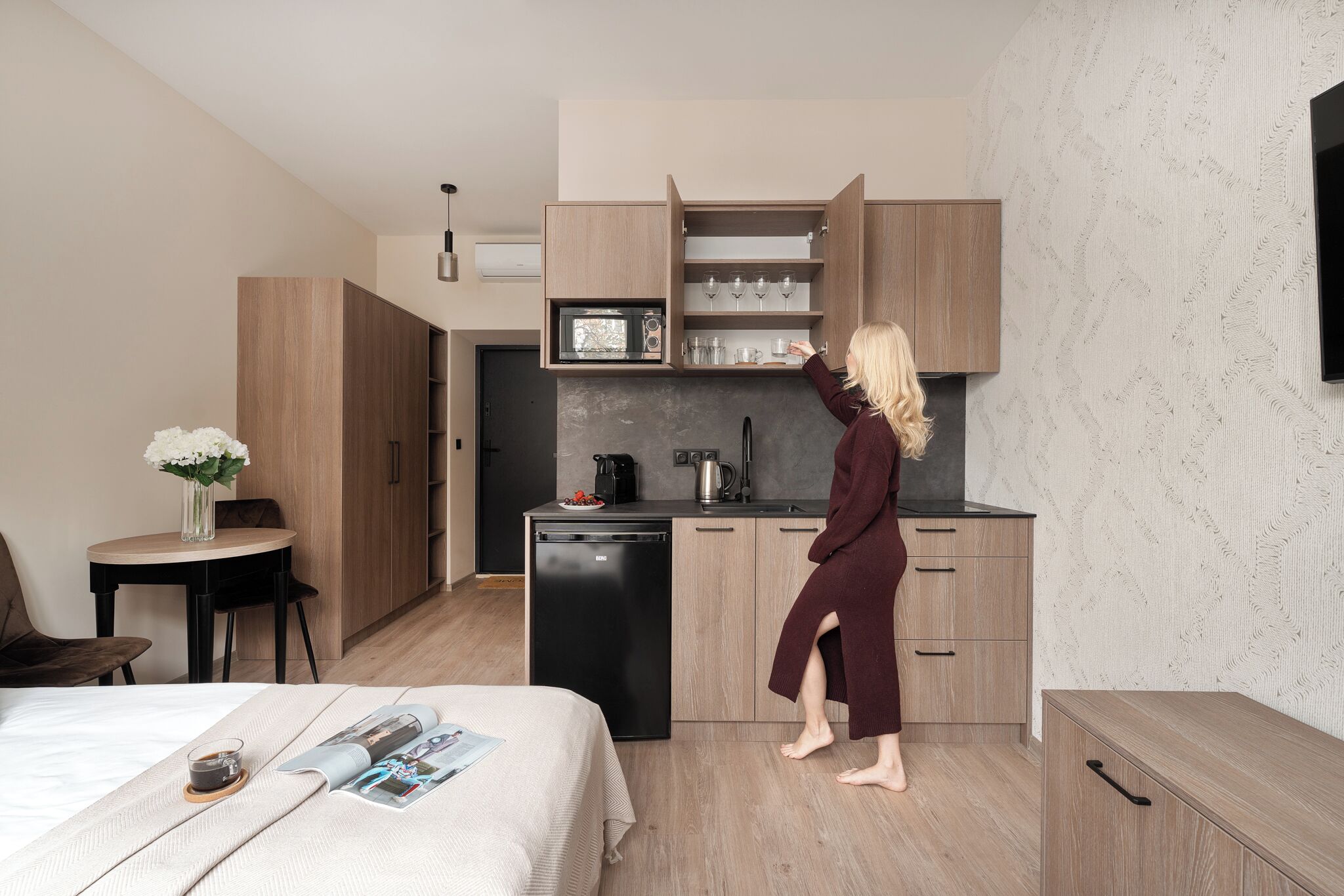 Habitación de hotel pequeña con kitchenette, una cama con una revista y una taza, y una mesita con un florero.
