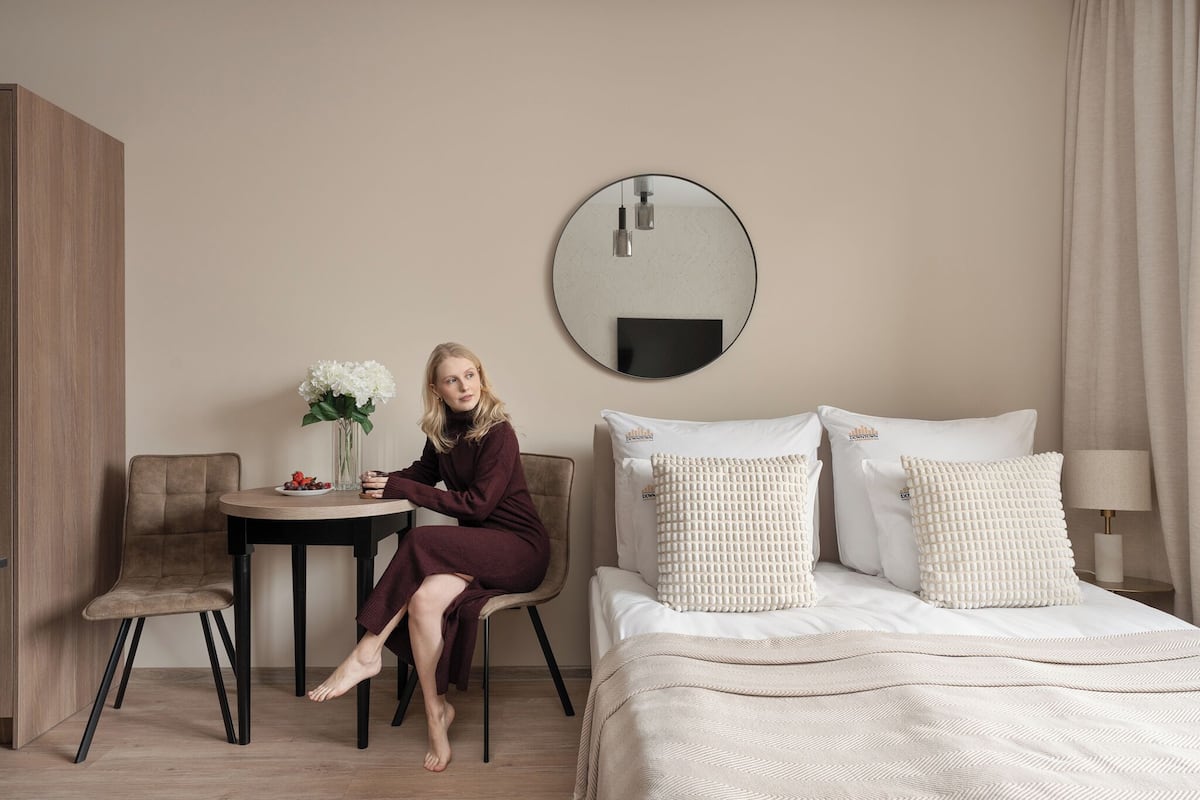 Habitación de hotel con cama, espejo redondo, una mesita con un jarrón de flores y una silla.