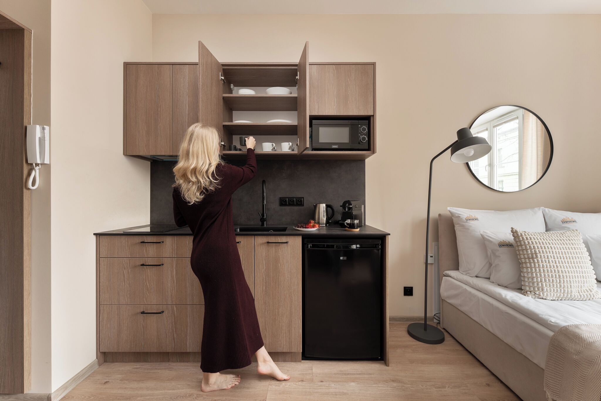 A modern kitchen with wooden cabinets, a microwave, and a coffee maker. There is a sofa with a mirror and a floor lamp in the adjacent living area.