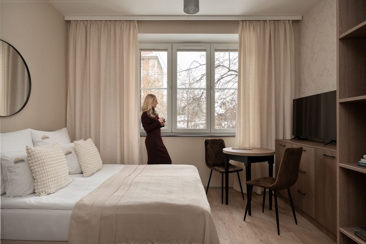 Habitación de hotel con cama, una silla, una mesita, televisión y ventana con cortinas.