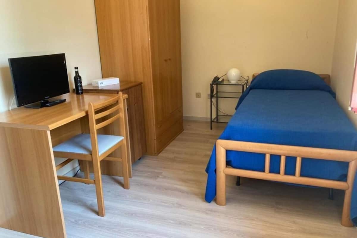 Un dormitorio con un escritorio de madera, una cama con colcha azul y una mesita de noche con lámpara.